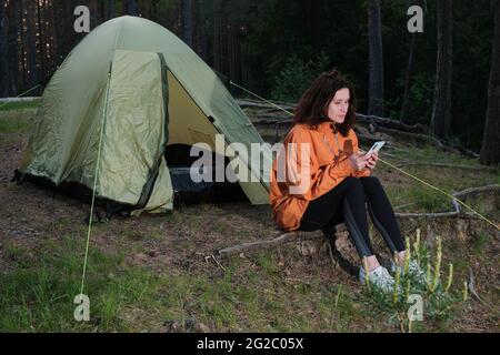 The girl sits next to the tent at night and chatting on the smartphone. Travel outside the city in the woods. Camping. Stock Photo