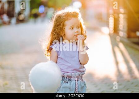 Little curly gir with white sweet cotton candy in the park at summer sunset Stock Photo