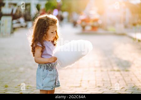 Little curly girl eat white sweet cotton candy in the park at summer sunset. Copyspace Stock Photo