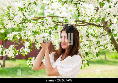 Young beautiful smiling pretty woman posing against the background of a blossoming apple tree. Spring lifestyle portrait. Cute happy brunette Caucasia Stock Photo