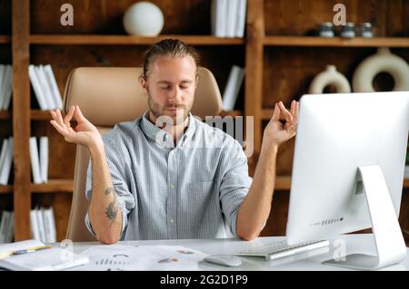 Relaxed peaceful young adult caucasian manager, businessman or office employee, sitting in office at table, taking break, freeing his head from thoughts, meditation, closing his eyes, calmness concept Stock Photo