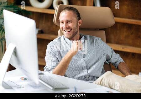 Happy confident attractive young adult manager or businessman, sitting in office with his legs on table, uses computer, communicates via video communication with colleagues or friends, smiles friendly Stock Photo