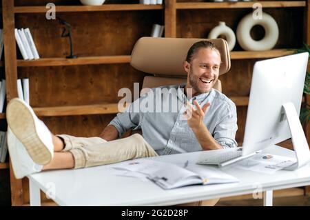 Joyful confident handsome young adult manager or businessman, sitting in office with feet on table, using computer, communicating via conference call with colleagues or friends, smiling Stock Photo
