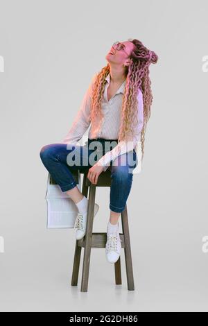 Woman with dreadlocks sits on tall chair, holds magazine in hand and looks up Stock Photo