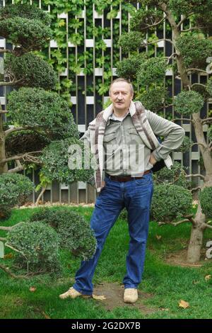 A white-haired fifty-year-old man in jeans and a shirt stands in a city park near a tree Stock Photo