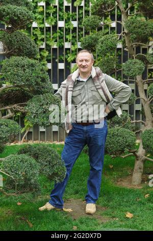 A white-haired fifty-year-old man in jeans and a shirt stands in city park near a tree and looks at camera with smile Stock Photo