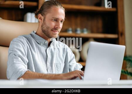 Successful smart focused young caucasian ceo, or business leader, sitting at work desk in office, using laptop, browsing internet, looking for ideas for project, answers e-mail, chatting online Stock Photo