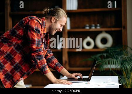 Attractive smart caucasian freelancer, using laptop. American stylish guy working remotely, stands near work desk, browsing internet, chatting online with colleague, smiling happily Stock Photo