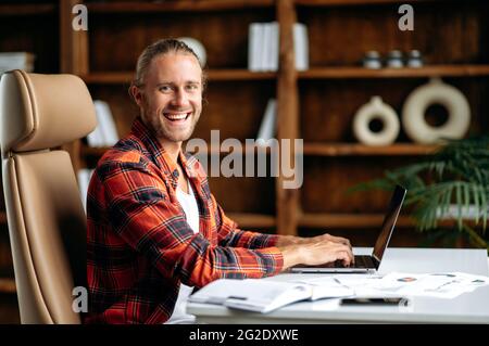 Portrait of joyful successful confident young adult caucasian freelancer or manager, using laptop. Handsome guy working distantly, sits at work desk, looking at the camera, smiling Stock Photo