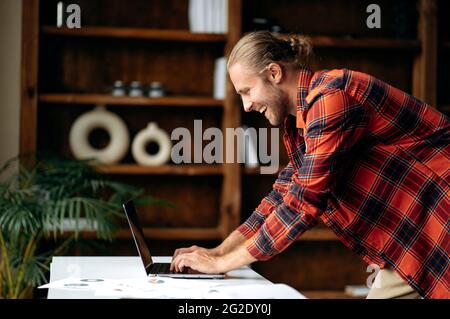Satisfied successful caucasian freelancer or manager, using laptop. American stylish guy working remotely, stands near work desk, browsing internet, chatting online with colleague, smiling Stock Photo