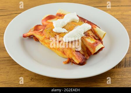 Three baked enchiladas with enchilada sauce and melted shredded cheese topped with sour cream on white plate Stock Photo