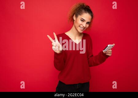 Photo of happy smiling woman with gathered curly hair wearing dark red sweater isolated over red background holding smartphone having fun and showing Stock Photo