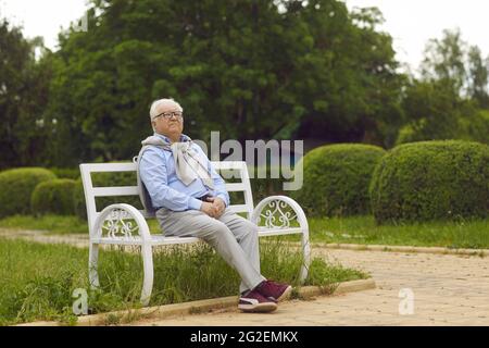 Senior man admiring nature while sitting on a white bench in a green summer park Stock Photo