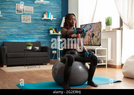African woman using stability ball keeping outstretched arms working out shoulders using blue dumbbells, in home living room for muscle shaping and healthy lifestyle, dressed in sportwear. Stock Photo