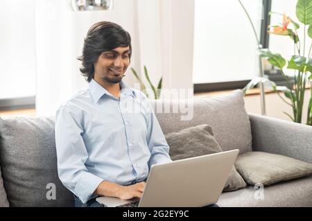 Happy indian freelance man using laptop for remote work from home, young man in casual wear sits on the sofa, texting, typing, writing an article, sending emails, chatting online Stock Photo
