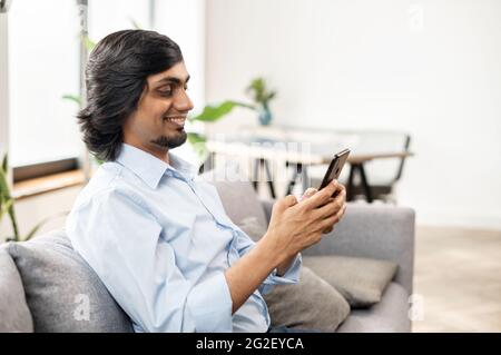 Serene young indian man holds smartphone sitting on the couch at home, eastern guy chatting online with friends, spend leisure time in social media, texting, scrolling news feed, shopping online Stock Photo