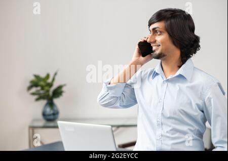 Smiling indian man, ceo, male employee in formal wear stands in bright office space and talking on the smartphone, looks away and has phone conversation Stock Photo