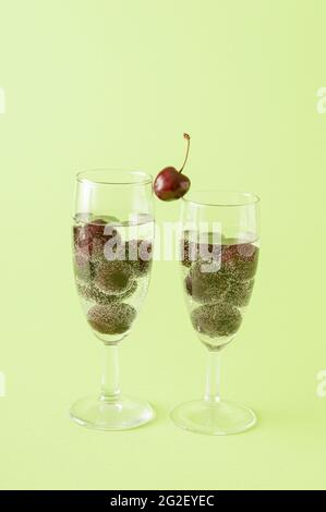 Two glasses of champagne filled with a cherry drink on a green background. One cherry between two glasses as a connection. Stock Photo