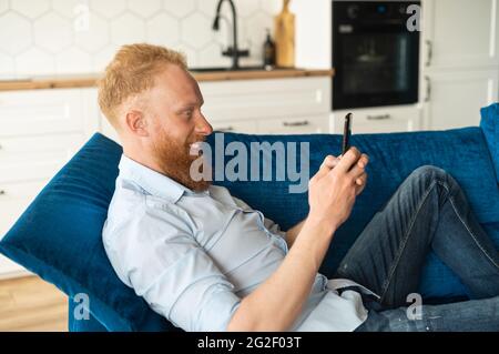 Serene smiling young redhead man lying down on the sofa at home with a smartphone and hands, checking emails, spending time in social networks, messaging and chatting online. Side view Stock Photo