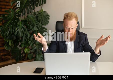 Perplexed and confused redhead bearded businessman staring at the laptop screen and makes a helpless gesture, a bearded hipster man in formal suit looking at monitor with asking expression Stock Photo