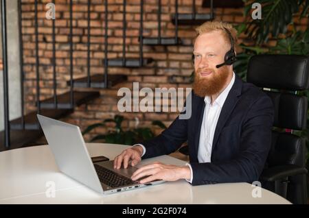 Inspired smiling young redhead businessman using wireless headset and laptop computer for online communication on the distance, cheerful optimistic businessman wearing headphones with mic looks away Stock Photo