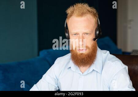 Young redhead man in smart casual shirt wearing wireless headset looks at the camera with serious face, close-up portrait of responsible support representative indoor. Diversity in work team Stock Photo