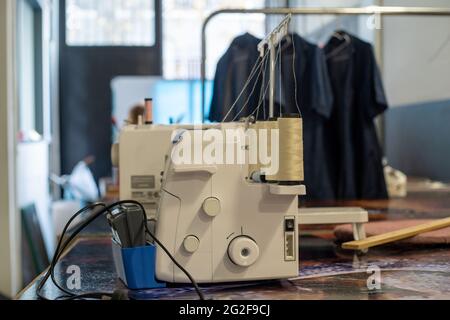 Closeup of overlock sewing machine on the table with hanged black coats on the rack behind it Stock Photo