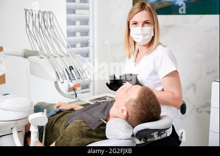 Horizontal snapshot of a pretty, young female dentist, wearing uniform, face mask, rubber gloves, sitting at her working place in well equipped dental office. Male patient during dentist's visit Stock Photo