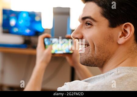 Happy brunette guy playing video game on his mobile phone at home Stock Photo