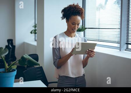 Mixed race businesswoman standing next to window, smiling and using tablet Stock Photo