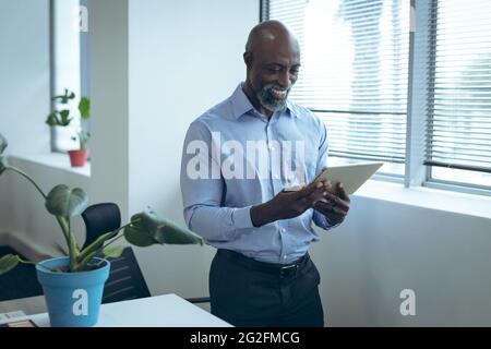 African american businessman standing next to window, smiling and using tablet Stock Photo