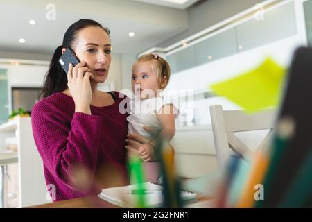 Caucasian mother holding her baby talking on smartphone while working from home Stock Photo