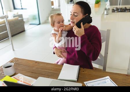 Caucasian mother holding her baby talking on smartphone while working from home Stock Photo