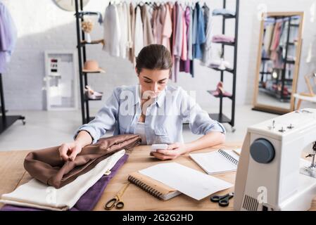 Seamstress using smartphone near cloth, notebooks and sewing machine Stock Photo