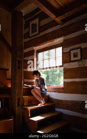 Boy reading a book indoors on the sunny step of a log cabin home. Stock Photo