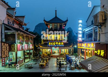 Night view of a square with table and chairs surrounded by illuminated traditional Chinese restaurants against a mountain backdrop, Yangshou, China Stock Photo