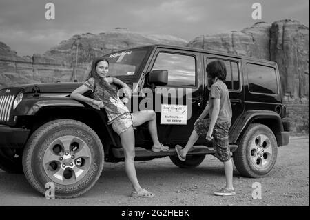 A girl and a boy in front of a Jeep Wrangler and a classic view of Monument Valley from Artist Point. Monument Valley Navajo Tribal Park, Utah and Arizona, USA, Stock Photo