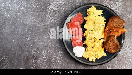 Scrambled eggs with salmon and toast on a round plate on a dark background. Top view, flat lay Stock Photo