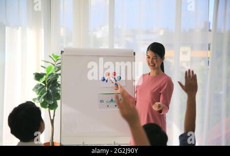 Smiling millennial female coach or presenter talk interact with multiracial colleagues at meeting, happy young woman tutor make flip chart whiteboard Stock Photo