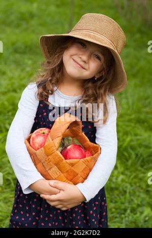 Happy Joyful Little Six Year Old Girl In Wicker Hat Hold In Her Hands Wicker Basket With Red Ripe Apples In Garden Close Up. Little Girl With Red Appl Stock Photo