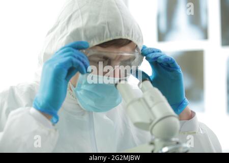 Doctor in a protective suit with gloves adjusts protective glasses near a microscope in a medical laboratory Stock Photo