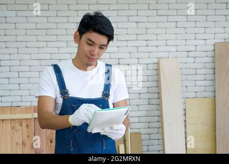 Asian carpenter inspect the list of items that must be done as scheduled, deliver the work with a happy face. Wood planks of various sizes lean on wal Stock Photo