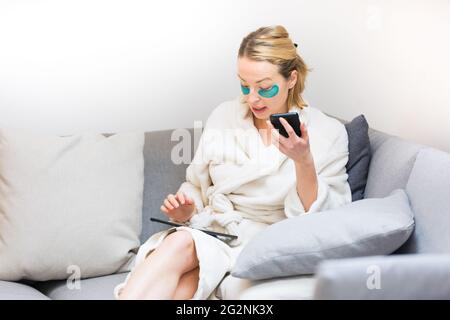 Young female entrepreneur wearing beauty face mask and cosy warm bathrobe in the morning, working remotly from her living room sofa. Remote work from Stock Photo