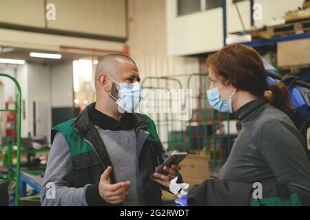 Industrial workers with face masks protected against corona virus discussing about production in factory. People working during COVID-19 pandemic. Stock Photo