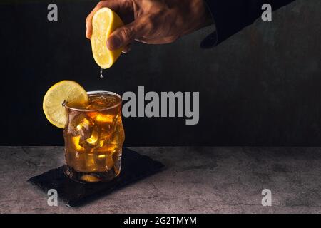 glass of tea with ice and lemon on stone surface with dark background and the hand of a man in a black shirt squeezing the juice of half a lemon on to Stock Photo