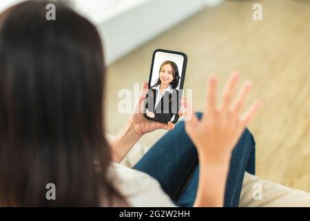 Young woman sitting on sofa holding mobile phone, making video call, discussing with company colleagues in telecommuting Stock Photo