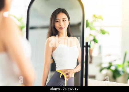 young woman takes a paper measuring ruler and measures her waist size before mirror. She is very satisfied with the results of the weight loss and the Stock Photo