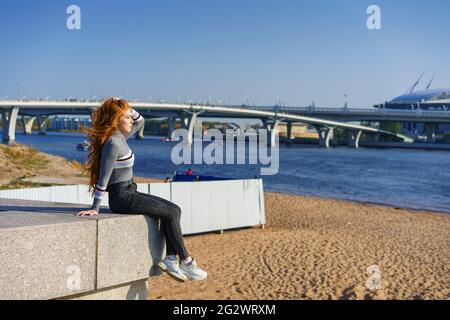 Beautiful young woman with long hair of Caucasian nationality , in casual clothes sits on the embankment of the river on a spring sunny day with a view of the bridge Stock Photo