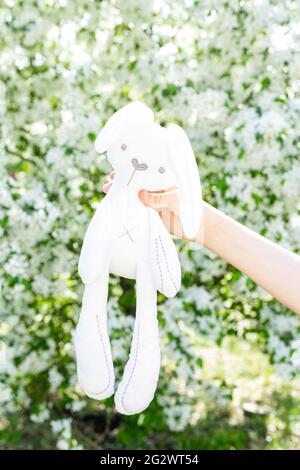 A child's hand holds a soft toy rabbit. A child on a walk in the park with his favorite soft toy. Bloomin apple tree background. Children's toys. Vert Stock Photo