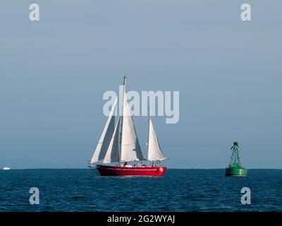 Classic style yacht with red hull sails past green buoy on beautiful deep blue sea. Stock Photo